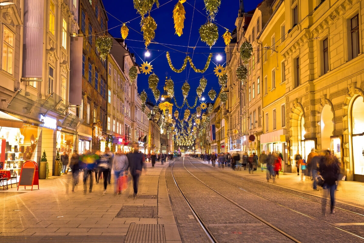 Karácsonyi fényekkel díszített Budapest utca estéje, ahol a lámpások és dekorációk meleg fényben ragyognak a téli vásár és városnézés közben.